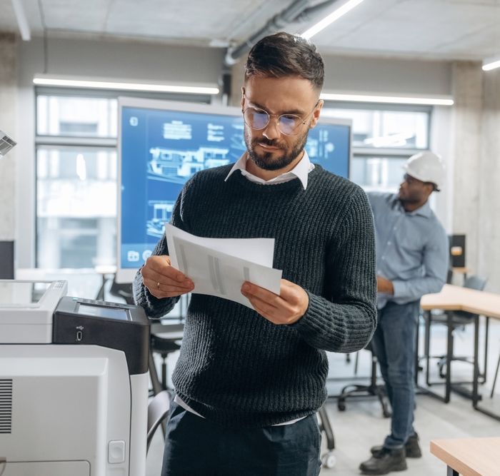 Person Standing Next to a Printer in an Office Environment Reading Documents