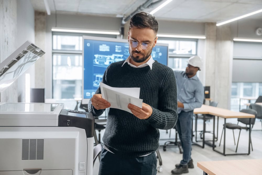 Person Standing Next to a Printer in an Office Environment Reading Documents